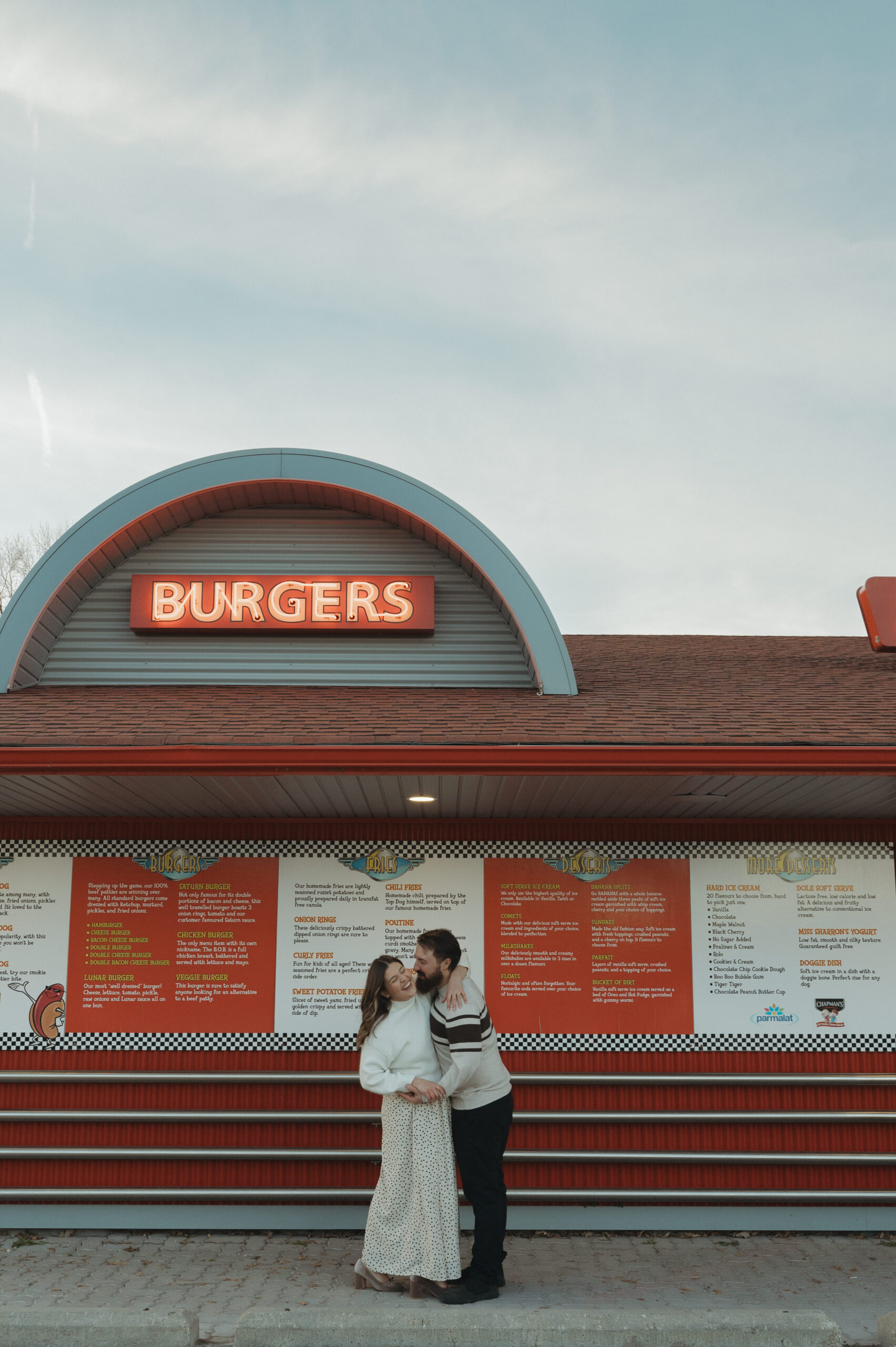 Couple giggle in front of an old retro diner sign for burgers, located in Manitoba for their engagement session